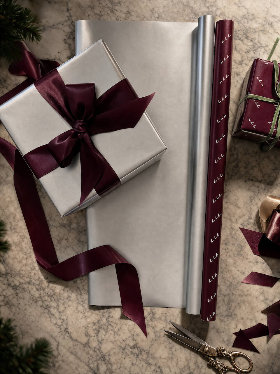 A styled flat-lay of wrapping paper products on a marble-like surface, including a silver gift box tied with a burgundy ribbon, a roll of silver wrapping paper, a roll of burgundy patterned wrapping paper, scissors, ribbon scraps, and another wrapped gift.
