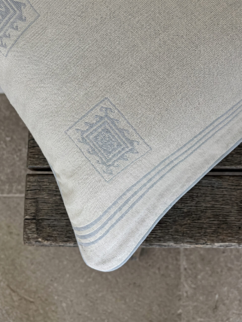 Close-up detail of a luxury cream and blue patterned BCI cotton cushion cover with woven geometric detailing and soft blue piping, styled on rustic timber for a relaxed contemporary aesthetic.