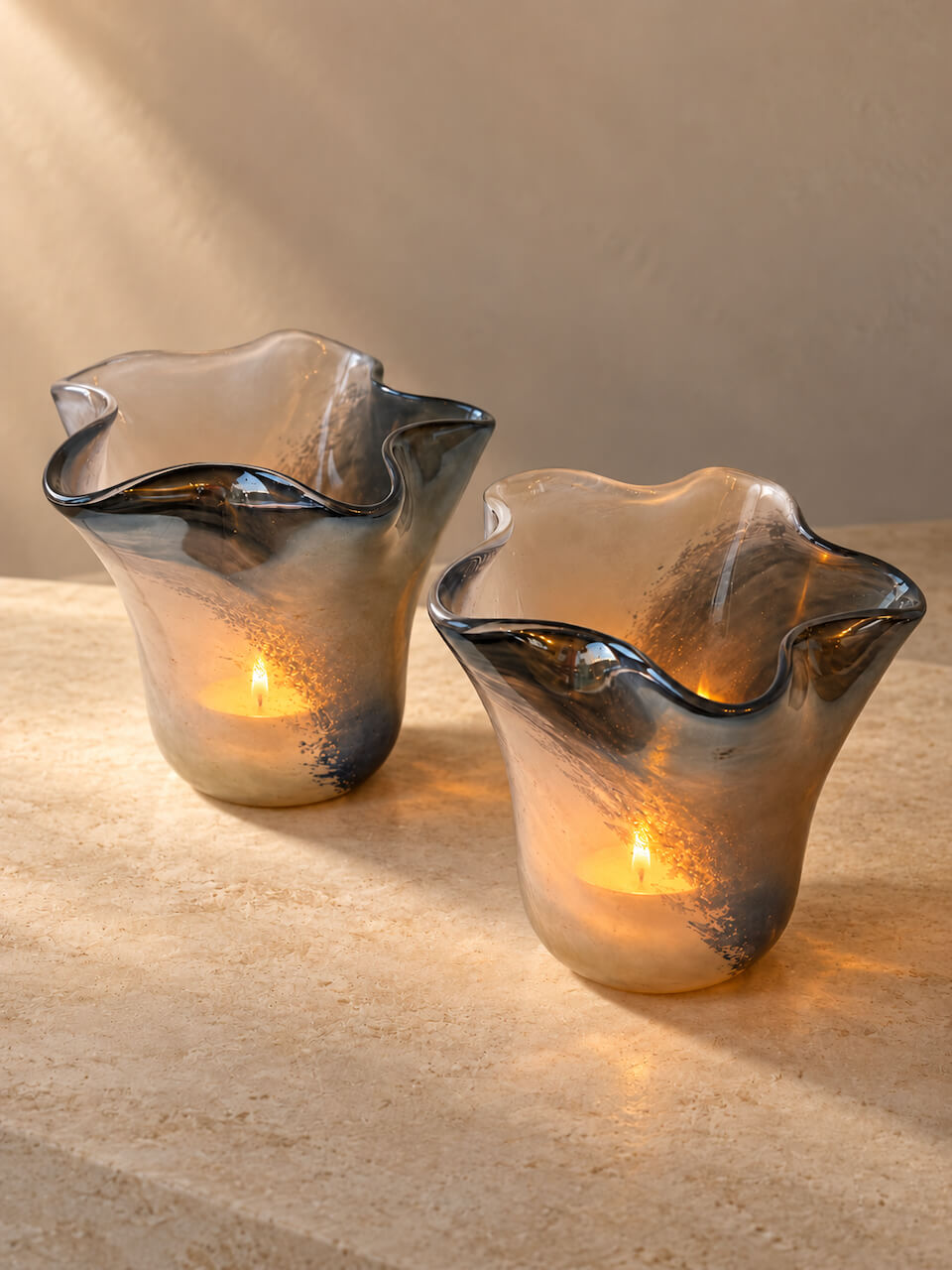 Two fluted glass candle holders with a smoky gray and black finish, each containing a lit tealight candle, set on a beige stone surface against a matching beige wall.