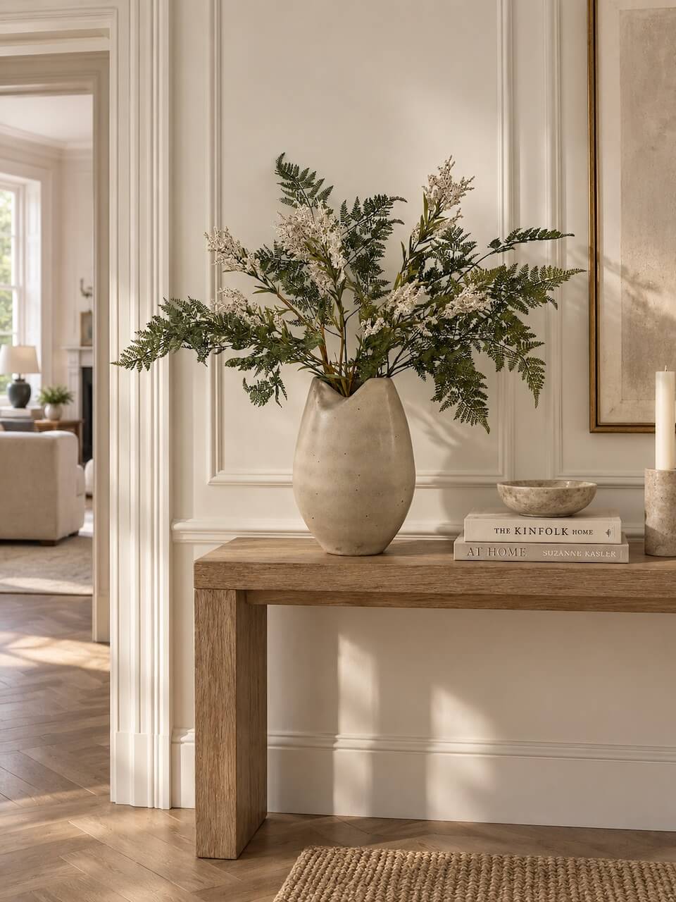 A tall textured beige ceramic vase filled with green and white dried botanicals, displayed on a rustic wooden console table in a bright, elegant room.