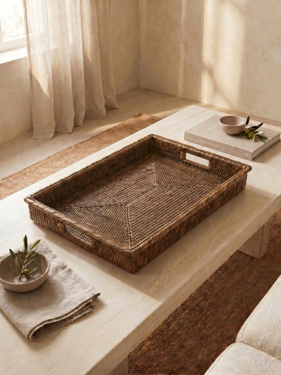 Large antique brown handwoven rattan tray with integrated handles styled on a light stone coffee table in a relaxed Mediterranean-inspired living space.