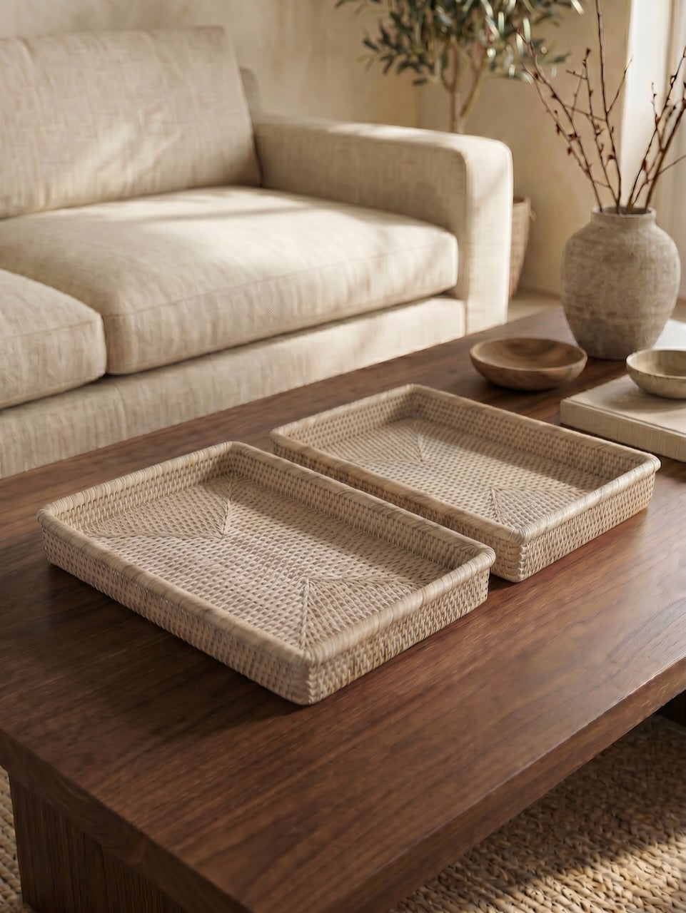 Two handwoven natural rattan trays styled side by side on a dark wooden coffee table in a relaxed neutral living room with warm sunlight and ceramic accessories.