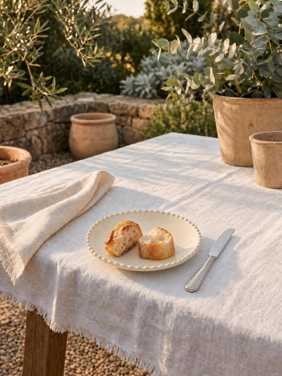 Cream scalloped side plate styled with rustic bread on a white linen tablecloth outdoors, surrounded by terracotta pots and Mediterranean garden greenery in warm evening light.