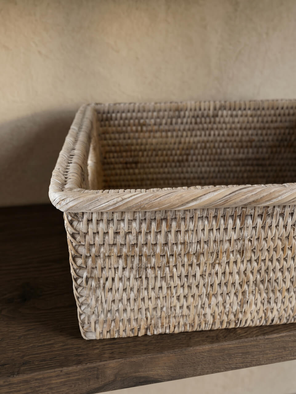 Close-up detail of a natural whitewash woven rattan storage basket with textured handcrafted weave, styled on a dark wooden shelf against a warm neutral wall.