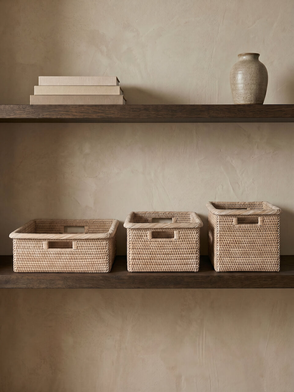 Set of three natural woven rattan storage baskets styled on dark wooden shelves against a warm neutral plaster wall, with minimalist decor and ceramic vase accents.