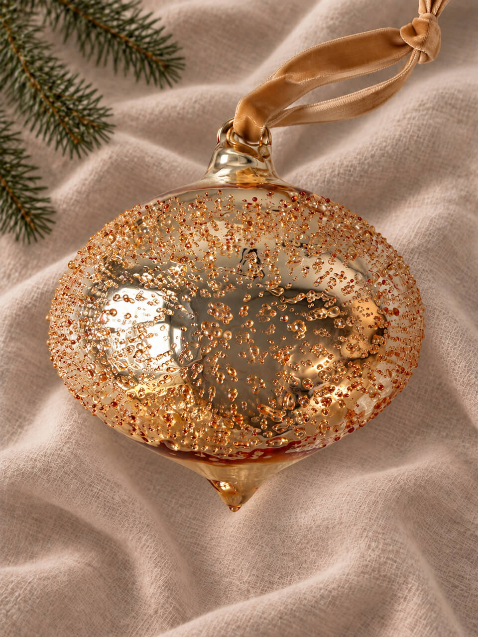 A gold glitter Christmas ornament with a hanging ribbon, styled on a beige linen fabric with pine branches in the corner.