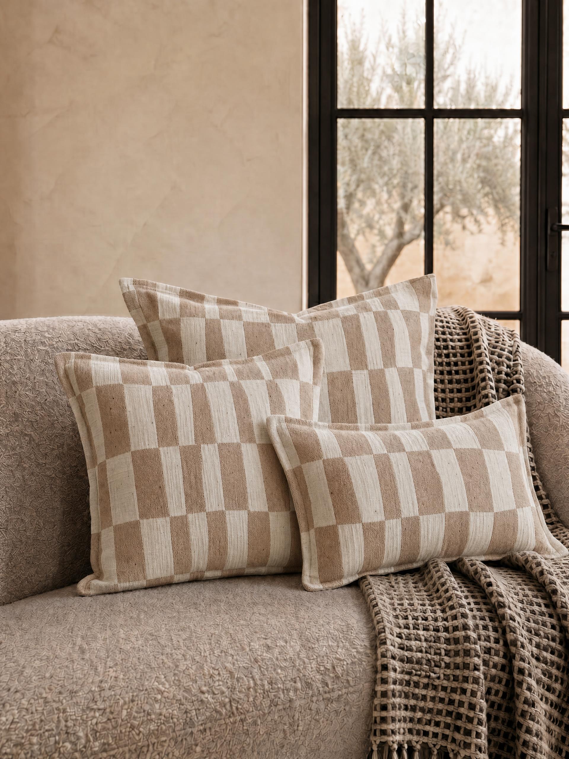 Three neutral woven check patterned cushions in layered sizes styled on a textured beige sofa with a taupe waffle throw and black framed window in a warm minimalist living room.