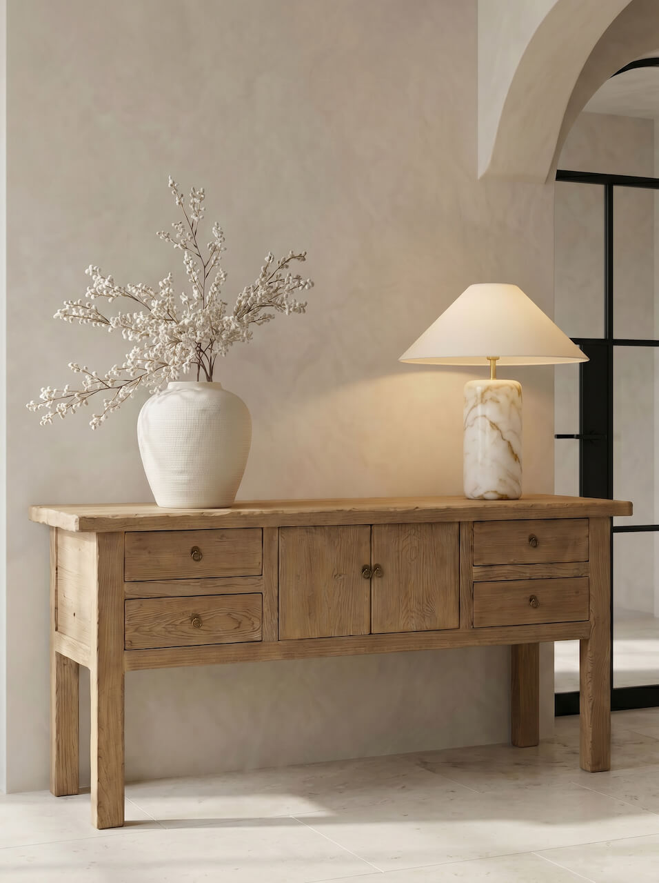 Wooden console table with a vase of flowers and a lamp against a beige wall.