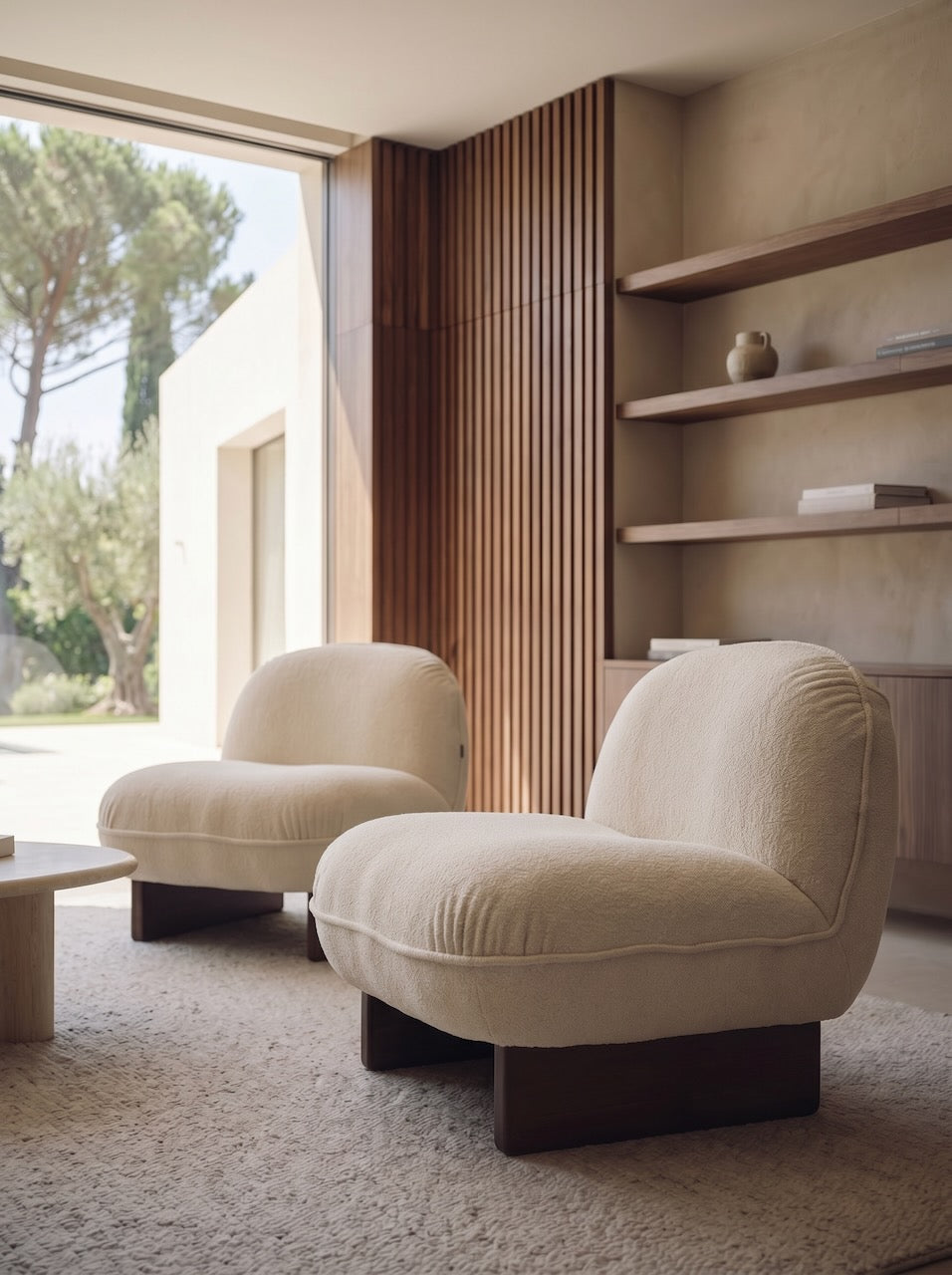 Beige armchair in a modern living room with large windows and wooden shelves.