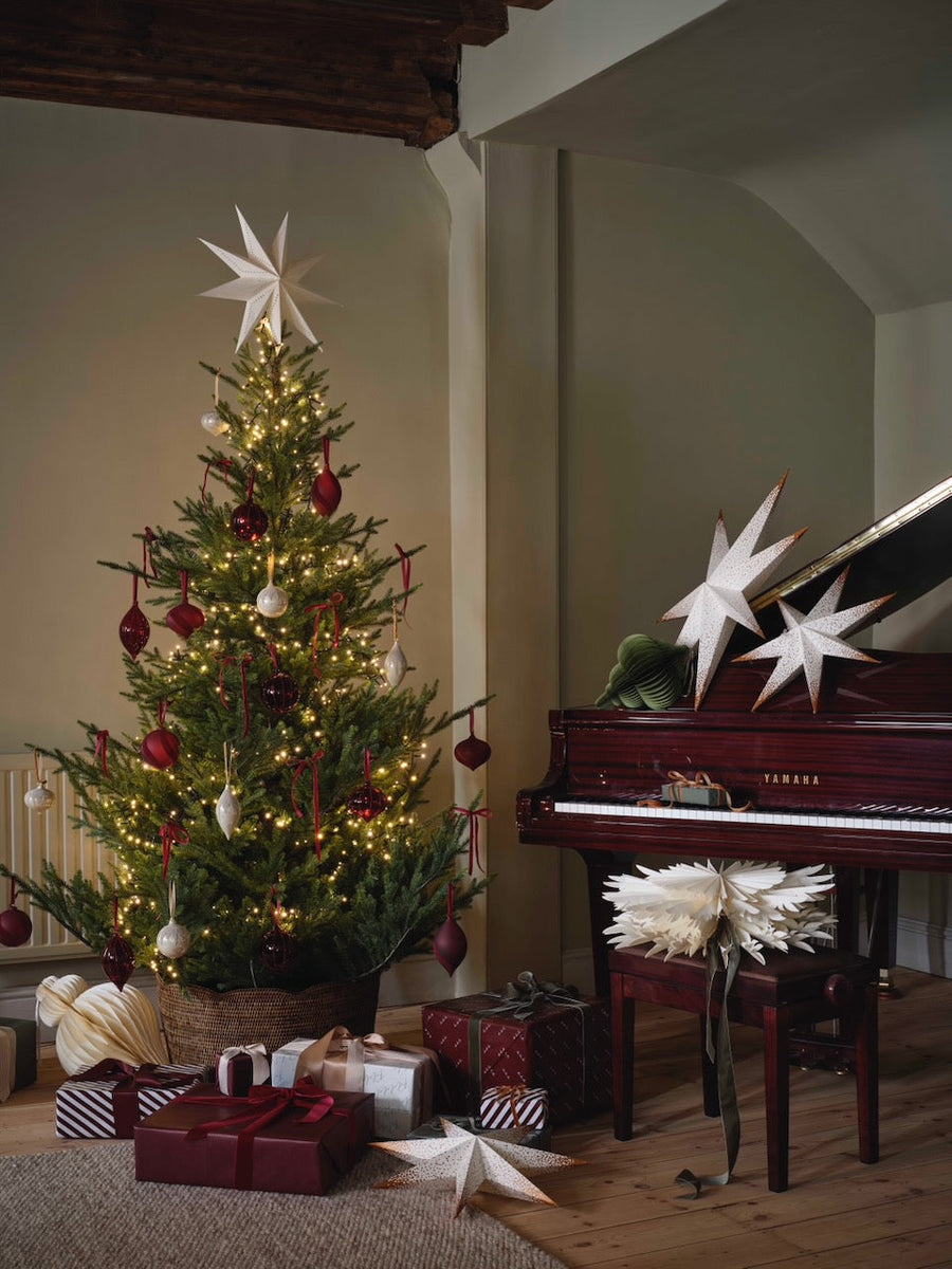 A decorated Christmas tree with warm white lights, red and white ornaments, and a large white star topper stands in a cozy room beside a dark red piano. Several wrapped gift boxes and white star decorations are arranged on the floor.