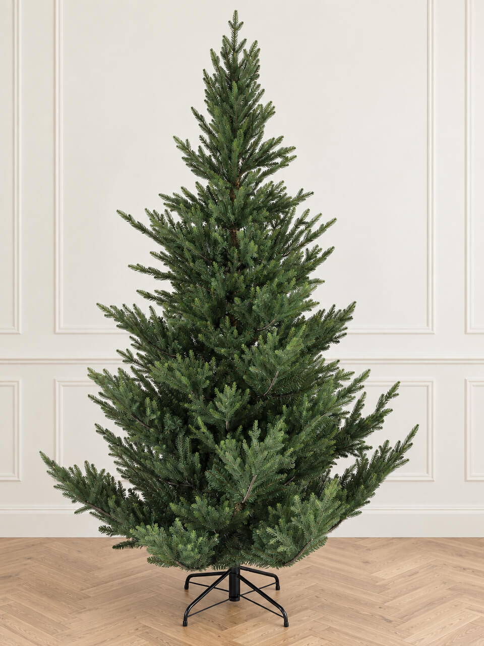 A tall artificial Christmas tree with dense green needles on a black metal stand, set against a cream paneled wall and light wood herringbone floor.