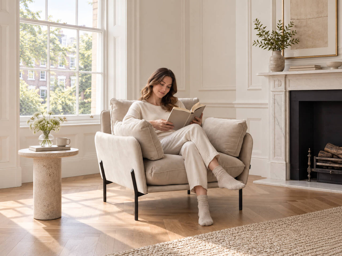 A woman in a beige lounge set sits in a cream upholstered armchair, reading a book in a bright, sunlit living room with a fireplace and wood floor.