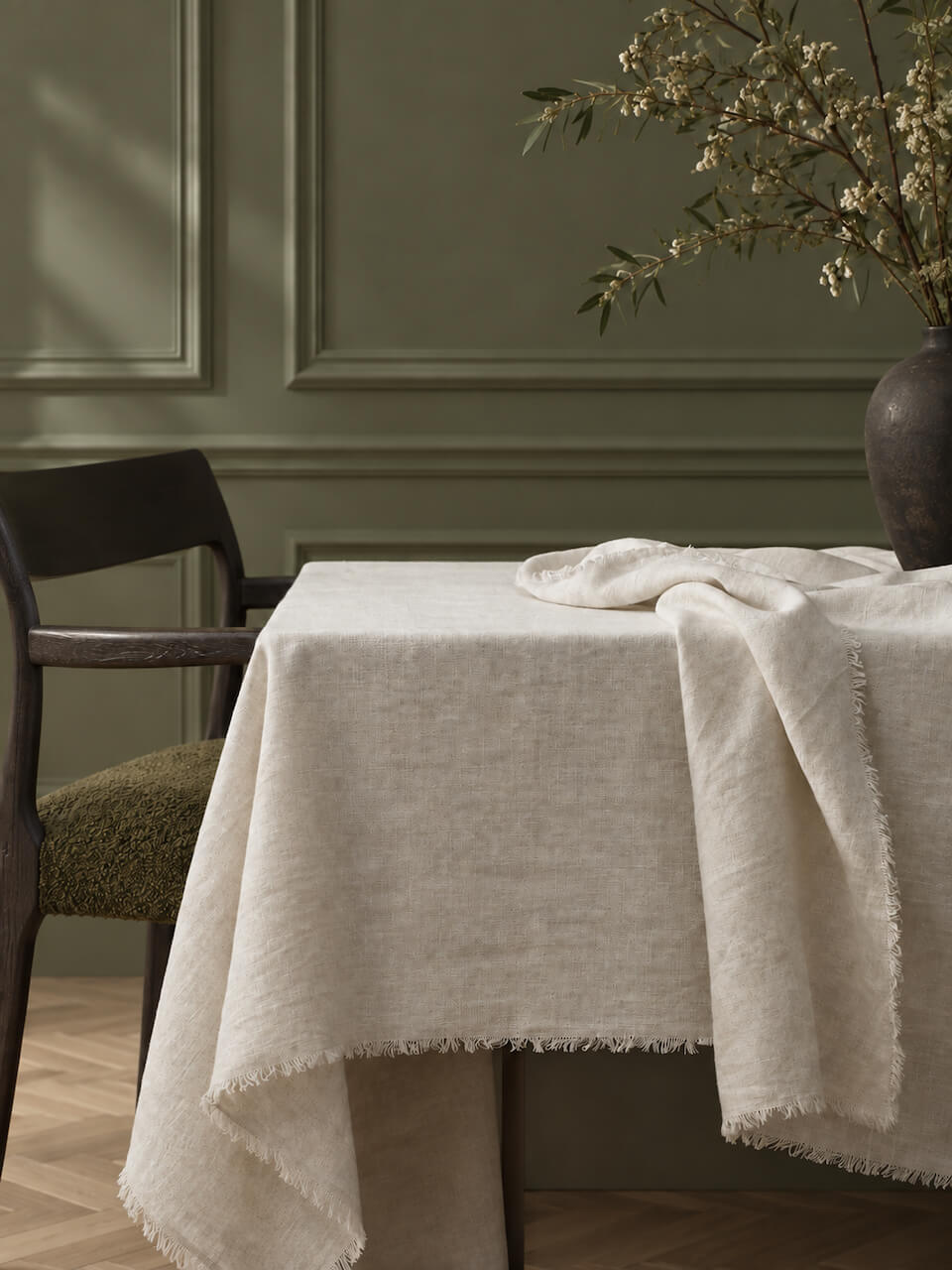 A antique-white coloured, fringed linen tablecloth draped over a table in a styled dining room setting with a dark wood chair and a vase of branches.
