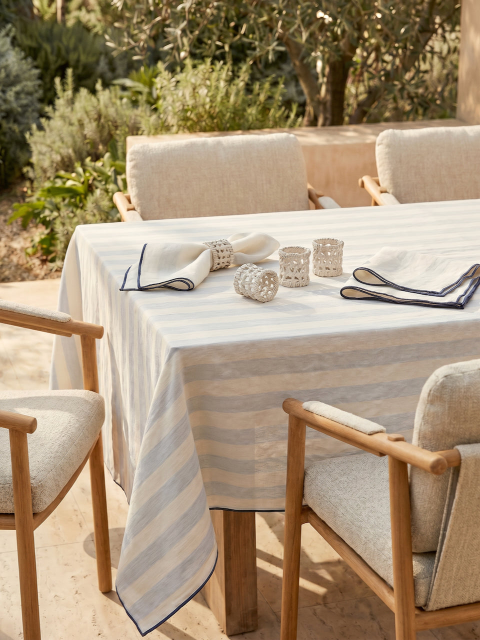 Set of four handwoven white rattan napkin rings styled on a soft striped linen tablecloth with neutral dining chairs in a sunlit outdoor Mediterranean dining setting.