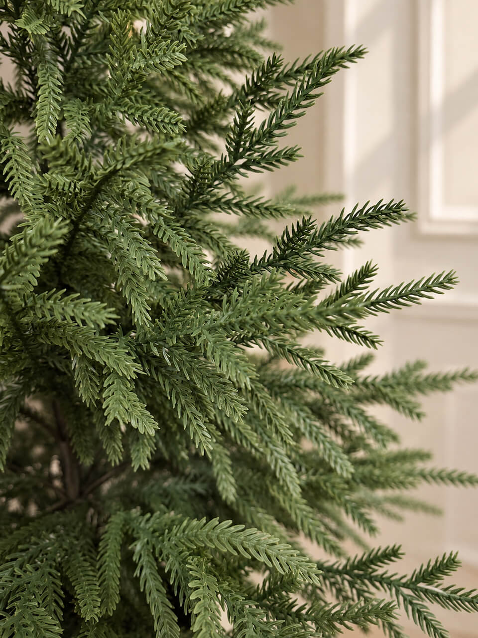 Close-up of green artificial pine branches with needle-like leaves, shown against a softly blurred cream-colored indoor background.