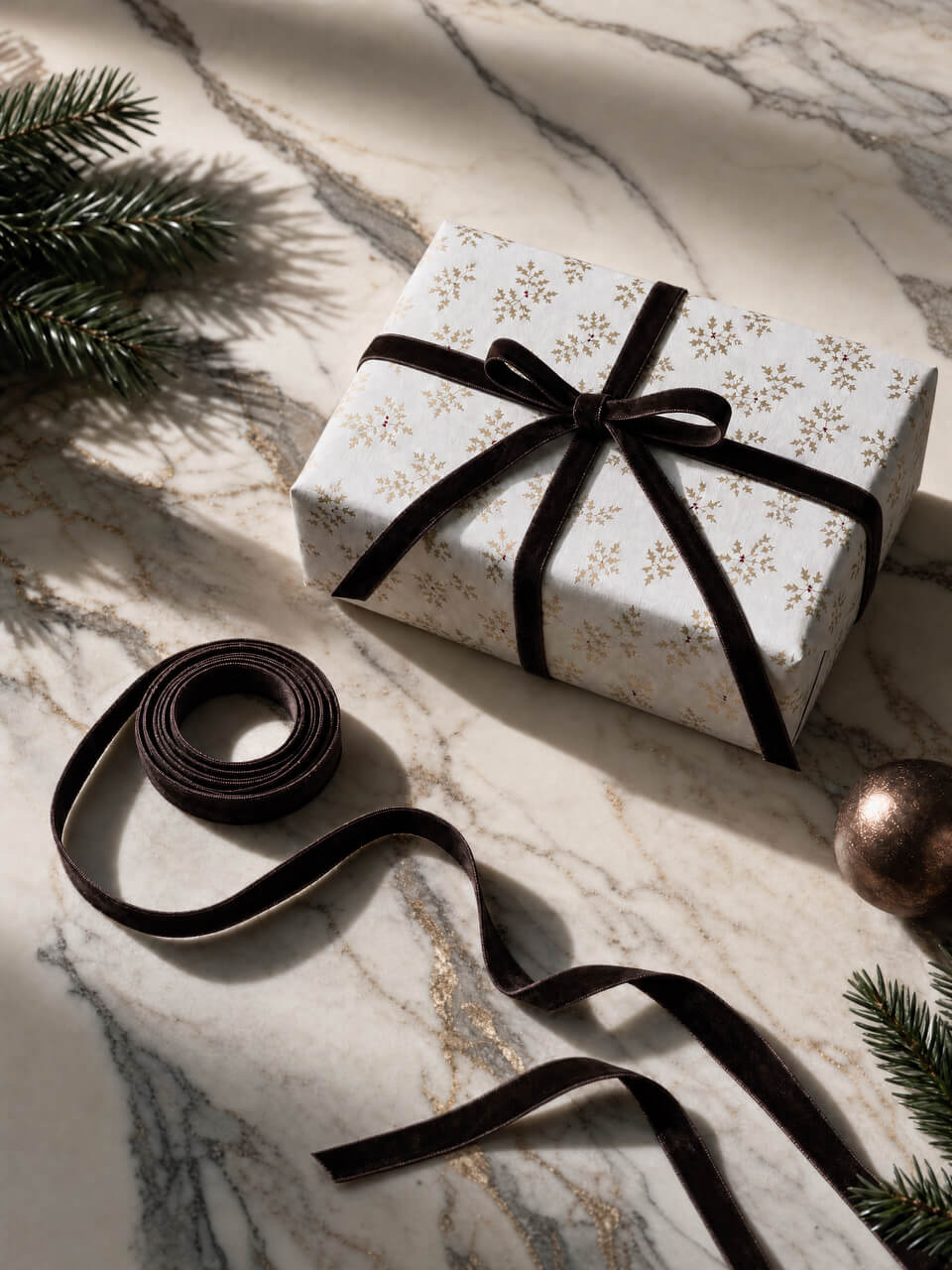 A wrapped gift box with white paper featuring a delicate gold snowflake pattern, tied with a wide black ribbon and bow, styled on a marble surface with pine branches and a metallic ornament.