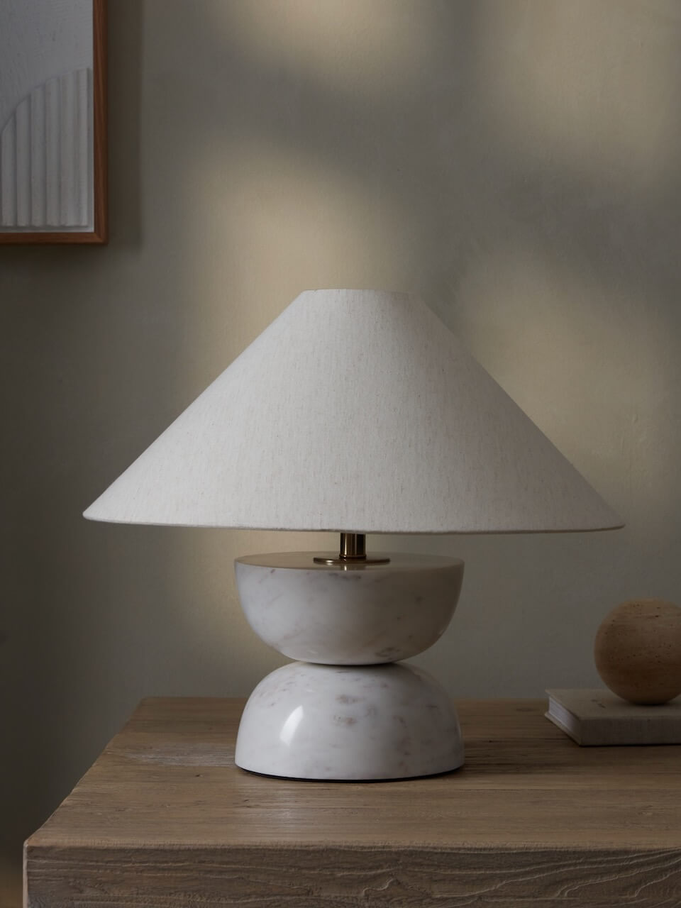 A white marble table lamp with a cone-shaped lampshade, placed on a wooden surface, with ambient lighting creating a warm tone on the wall.