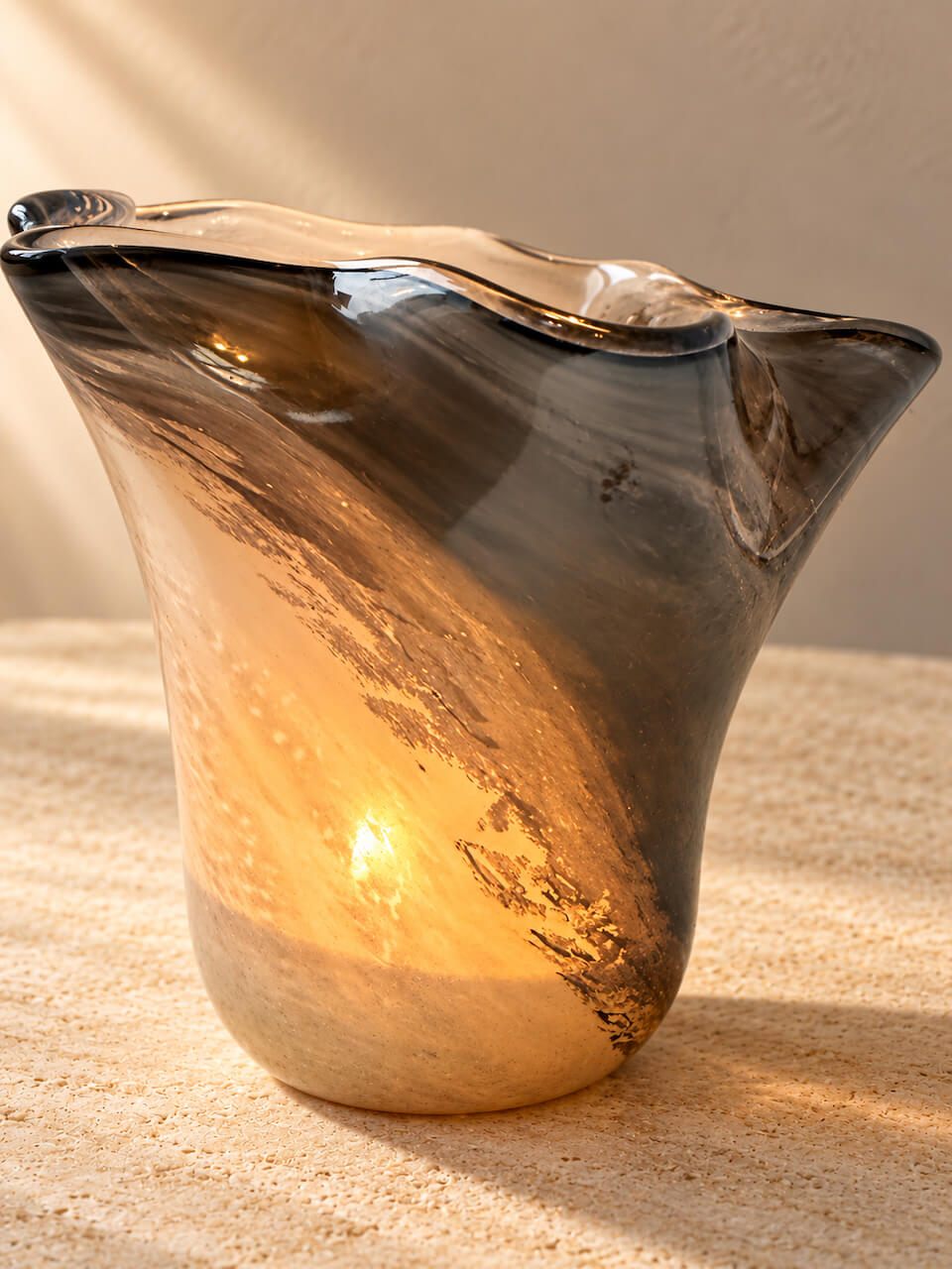 A sculptural, hand-blown glass vase with a wavy rim, featuring a gradient of dark gray and amber-brown glass that transitions into a warm, glowing golden-yellow interior, set against a neutral beige background.
