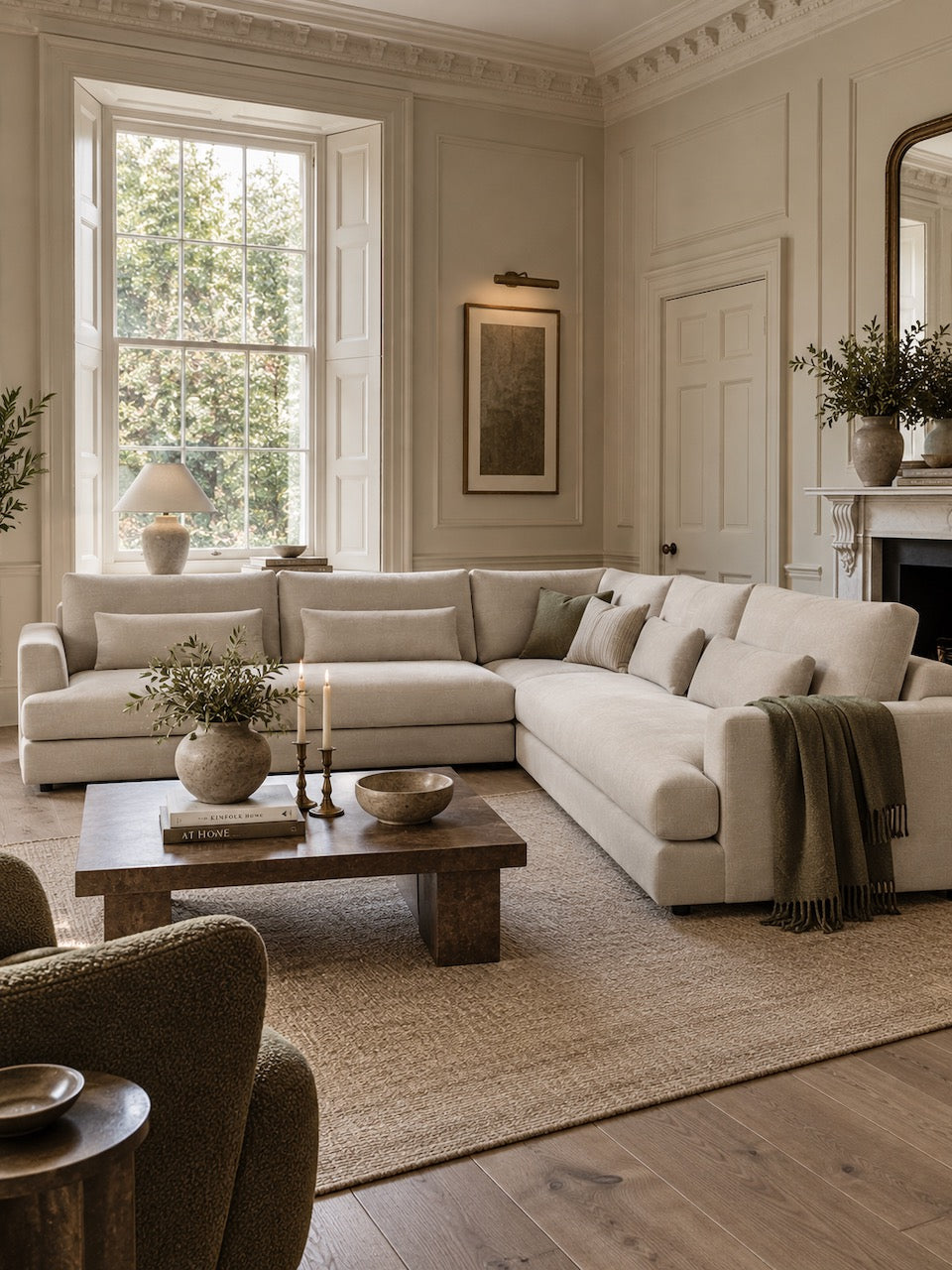 A large L-shaped sectional sofa upholstered in a light cream fabric, styled in a classic, elegant living room with a wooden coffee table, beige rug, and warm natural light from a bay window.