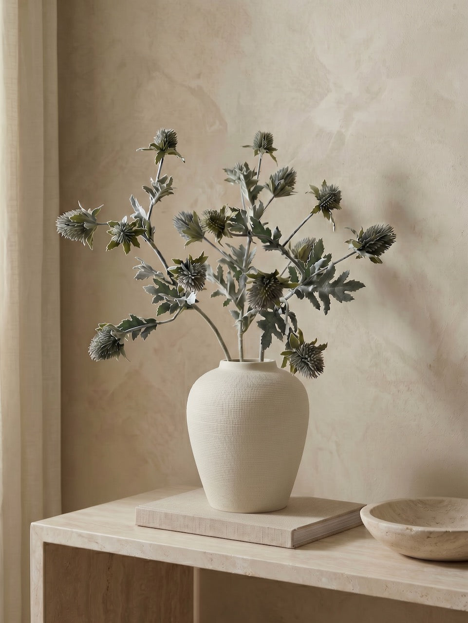 Cream Ceramic vase with faux thistle stems on a wooden table against a beige wall
