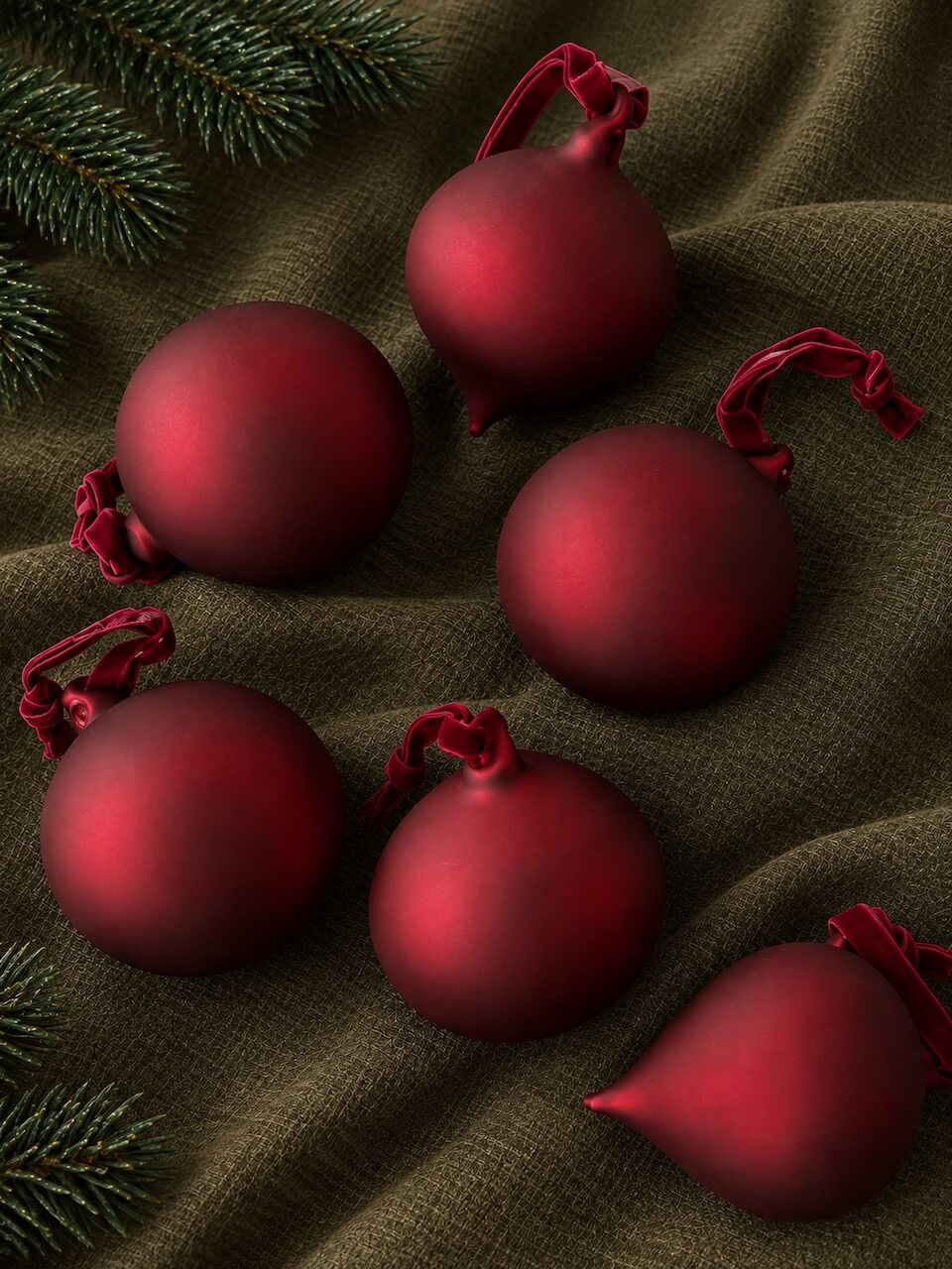Five matte burgundy Christmas baubles with red hanging loops arranged on dark olive green textured fabric with pine branches in the corners.
