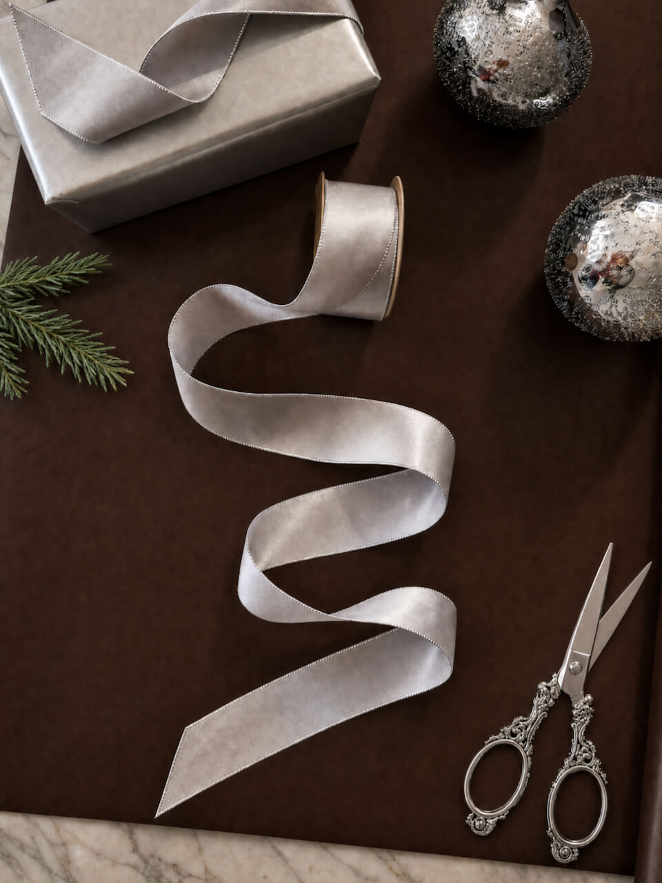 A spool of wide silver satin ribbon unrolled across a dark brown surface, with a wrapped gift box, silver ornaments, and ornate silver scissors arranged around it.