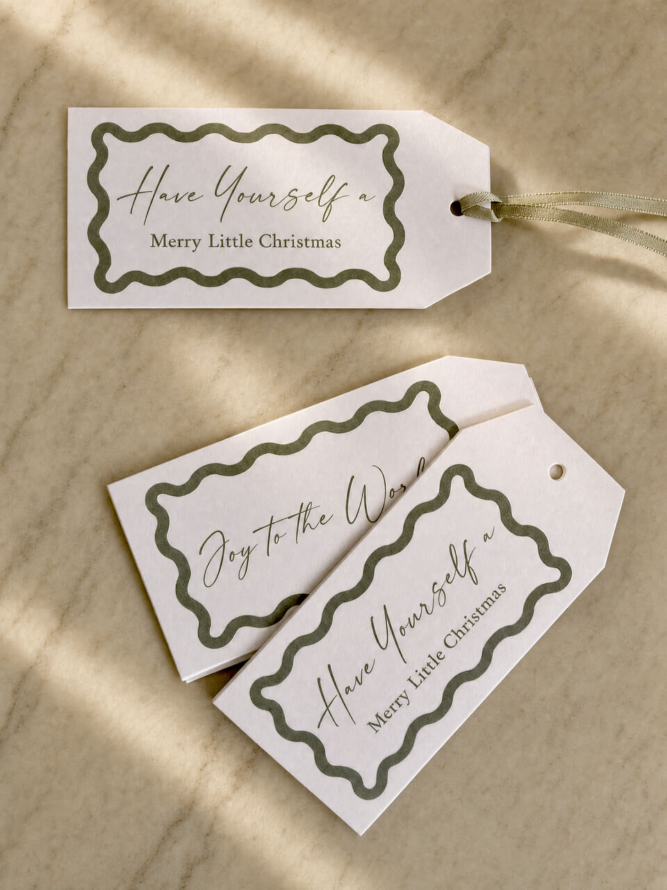 Three decorative gift tags with scalloped olive green borders and cursive holiday text, arranged on a light beige marble surface with soft shadows.