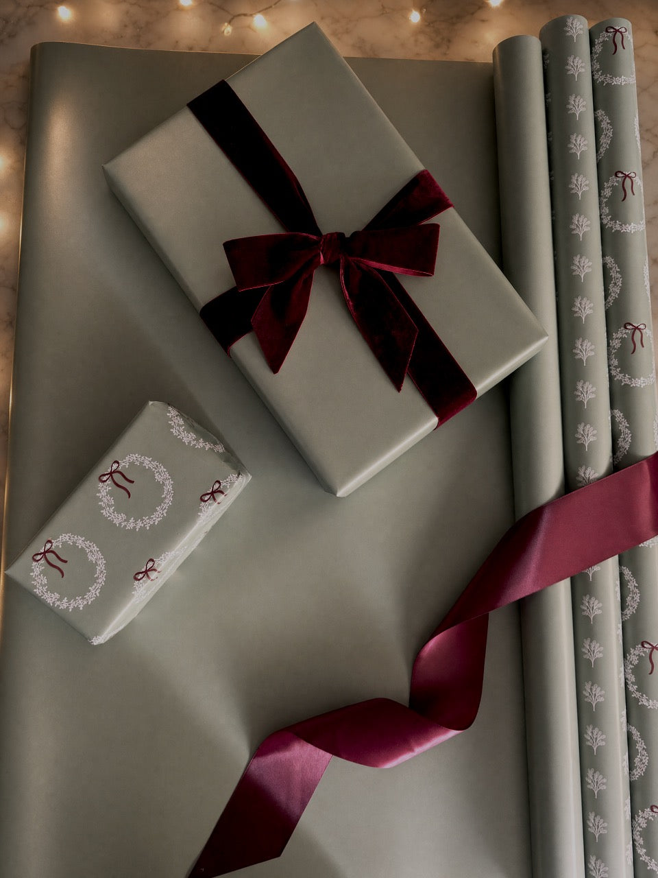 Gray wrapping paper with white wreath and bow patterns, shown wrapped around gift boxes and rolls with a dark burgundy ribbon on a neutral background.