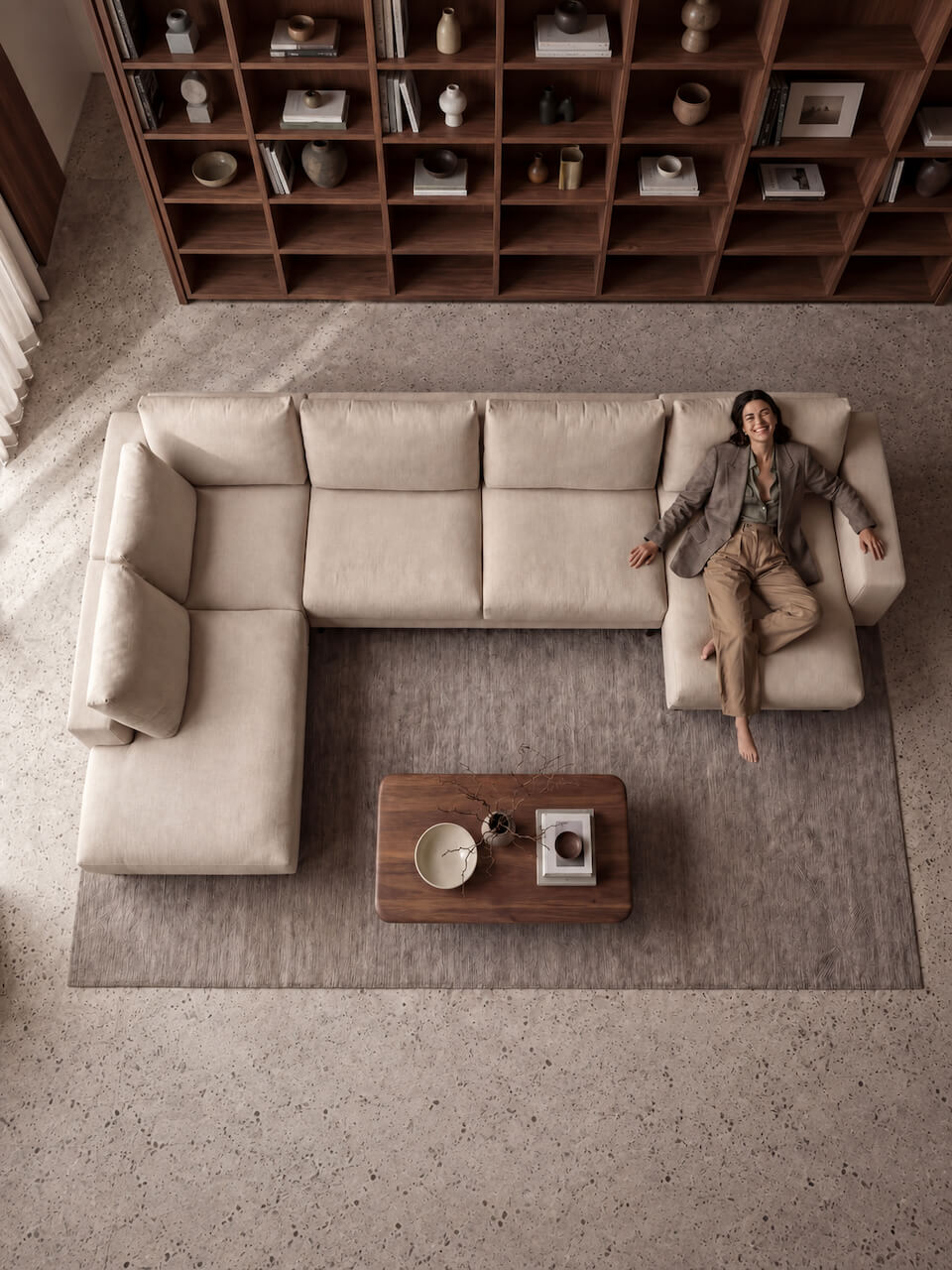 An overhead view of a beige sectional sofa with a person reclining on the right side, set in a modern living room with a wooden bookshelf and a coffee table.