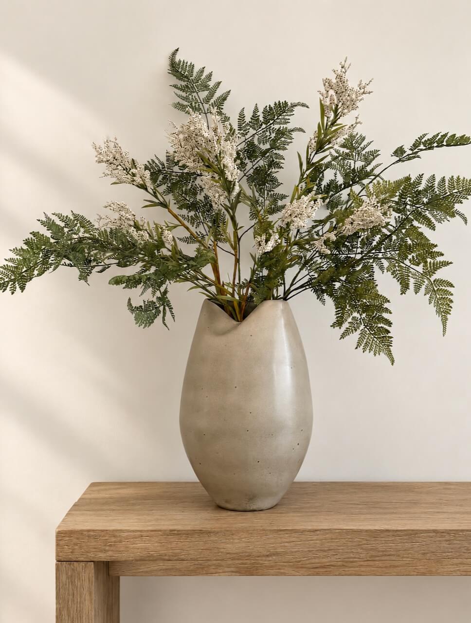 A matte beige ceramic vase filled with green fern-like foliage and small white dried flowers, displayed on a light wood table against a warm off-white wall.