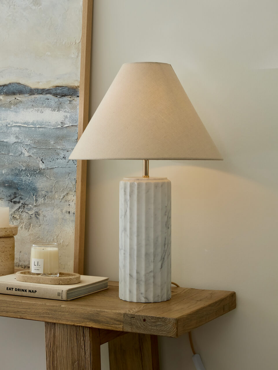 Table lamp with marble base on a wooden side table next to a candle and book, with abstract art in the background.