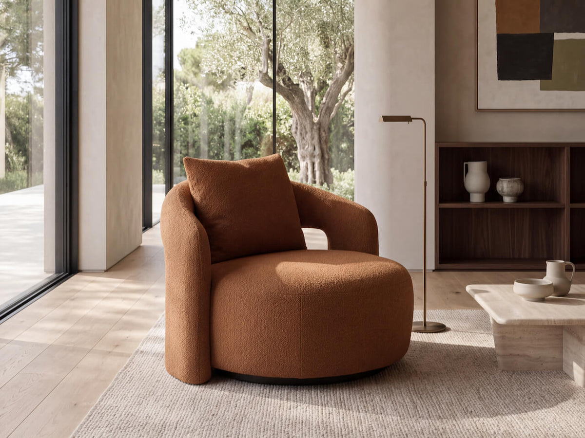 A modern round upholstered accent chair in a warm rust-brown fabric, styled in a contemporary living room with a matching pillow, a marble coffee table, and a wooden shelving unit in the background.