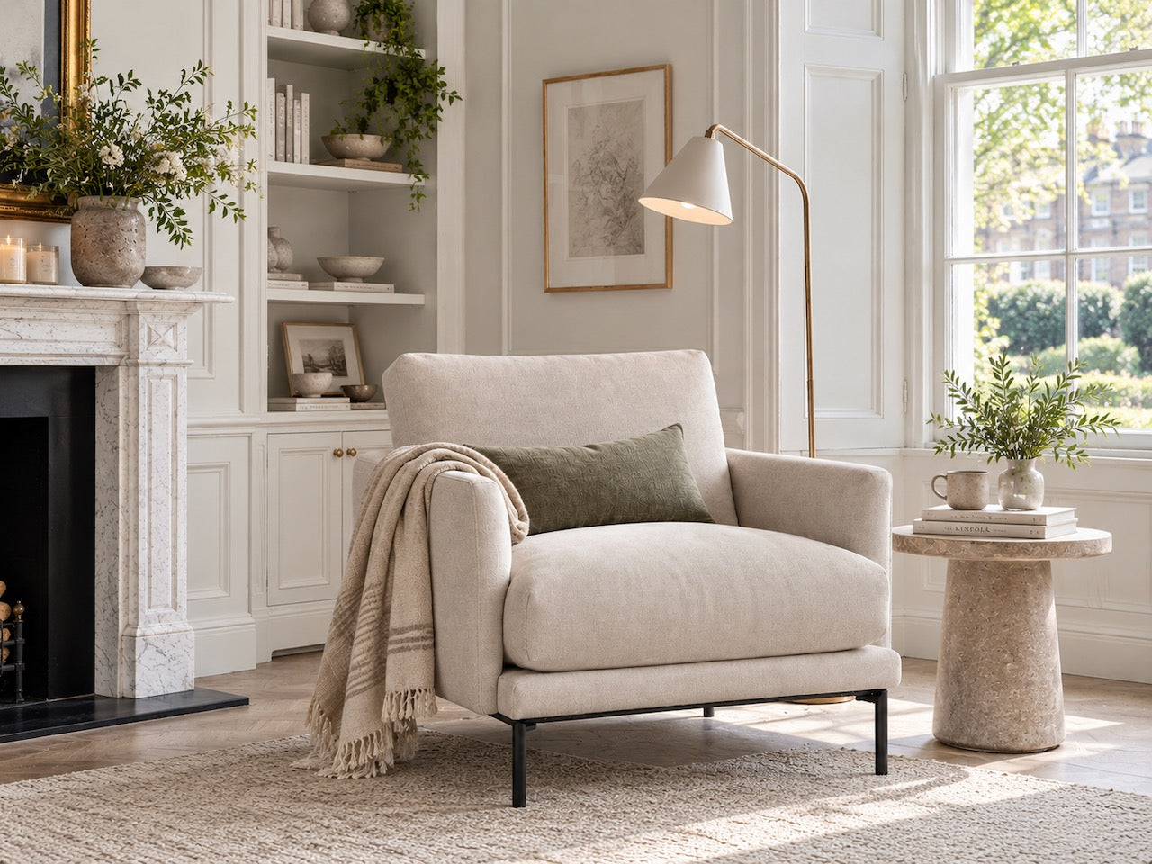 A light beige upholstered armchair with black metal legs in a bright, modern living room. A textured cream throw blanket and a dark green pillow are draped on the chair, which is positioned next to a stone side table, a floor lamp, and a white fireplace with built-in shelves.