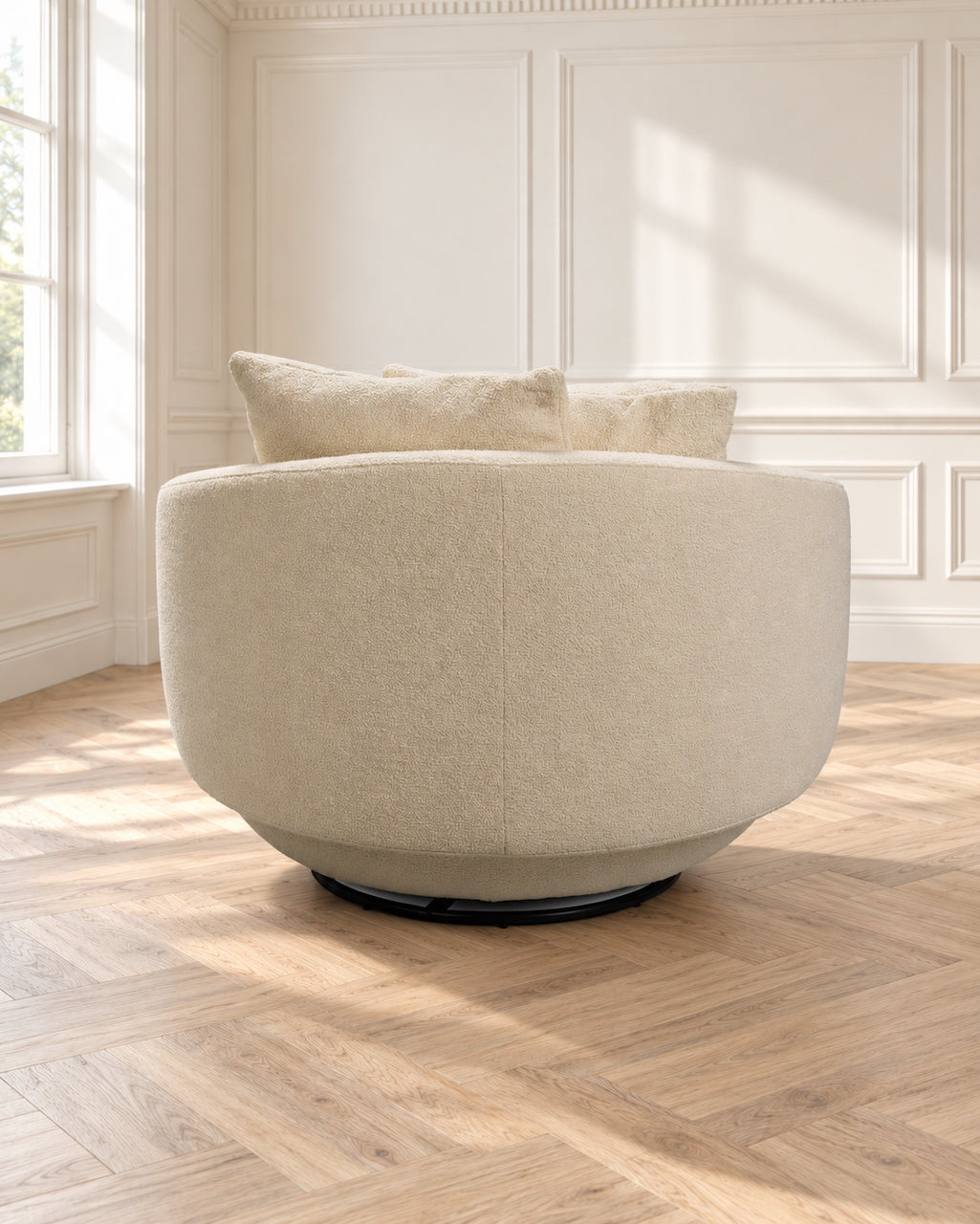 A round upholstered swivel armchair in a warm cream or beige fabric, shown in a bright room with white paneled walls and a light wood herringbone floor. Two matching throw pillows rest on the chair, and natural light streams in from a window on the left.