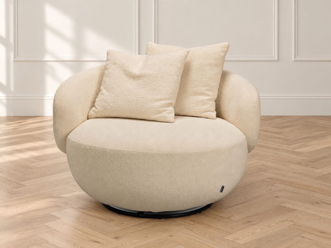 A modern round swivel armchair upholstered in a textured cream fabric, featuring two matching throw pillows and a black circular base, set against a light paneled wall and herringbone wood floor.