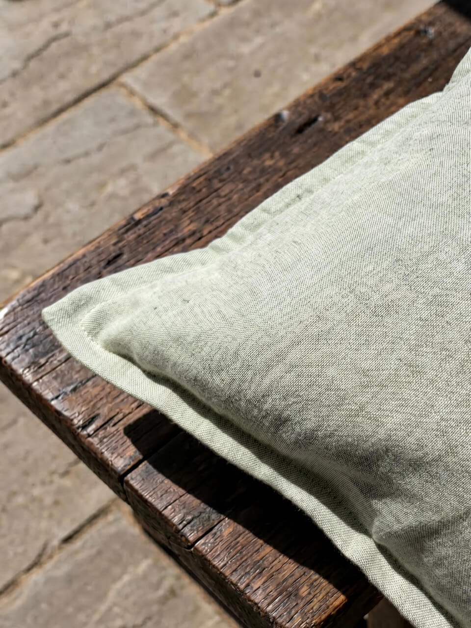 Close-up detail of a luxury sage green cotton linen cushion cover featuring a soft woven texture and tailored edging, styled on rustic timber in natural sunlight for a relaxed contemporary aesthetic.