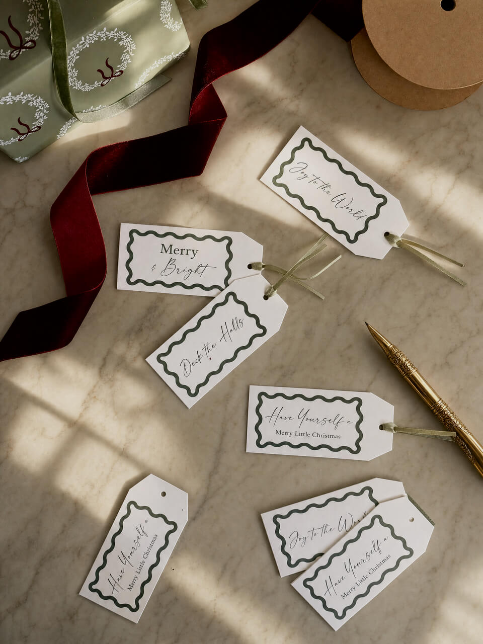 A styled flat-lay of several white gift tags with black decorative borders and cursive holiday messages, arranged on a marble surface with a dark red ribbon, kraft paper spool, gold pen, and wrapped gift box.