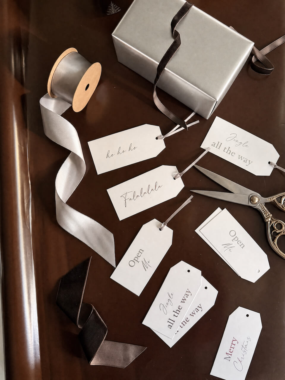 A styled flat-lay of gift wrapping accessories on a dark wooden surface, including a silver gift box with a dark ribbon, a spool of white ribbon, scissors, and multiple white gift tags with elegant script text.