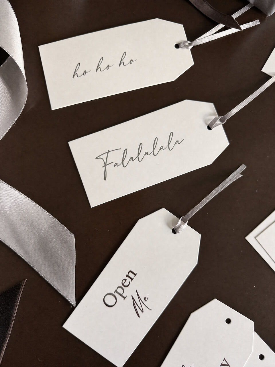 White paper gift tags with silver-gray script text, arranged on a dark brown background with silver ribbons.