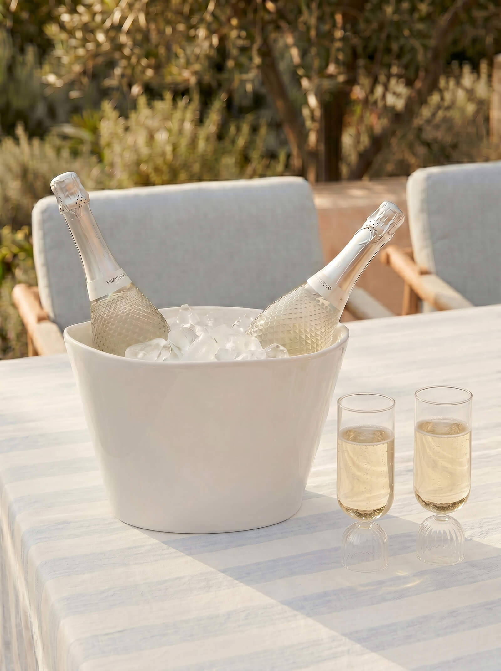 Two bottles of champagne in an ice bucket on a table outdoors with glasses of champagne.