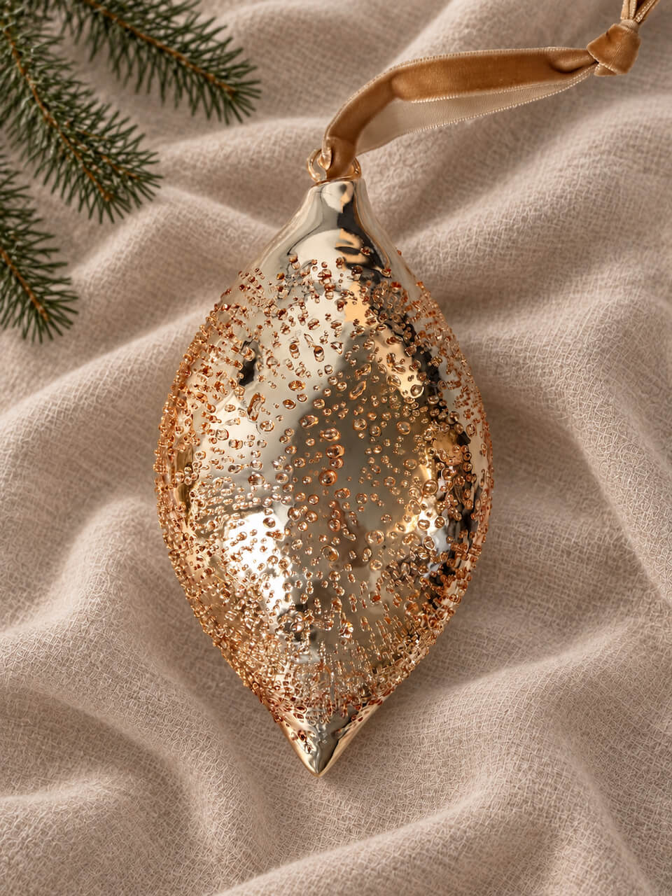 A teardrop-shaped holiday ornament with a metallic rose gold glitter finish and a matching ribbon hanger, displayed on a textured beige fabric with pine branches in the corner.